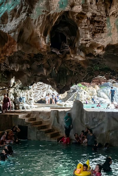 The image depicts a group of people bathing in a cave pool, possibly at the Wisata Alam Lumban Binanga in Penungkiren, Indonesia. The cave ceiling is adorned with stalactites, and natural rock formations create an enclosed environment. Several individuals are submerged in the clear, blue water, with some sitting on ledges and steps carved into the rock. A few people are standing or sitting on the platforms surrounding the pool. A bright yellow inflatable duck toy is visible in the foreground. The overall scene conveys a sense of recreation and natural exploration within the cave.