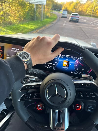 The image depicts a first-person view from the driver's seat of a Mercedes-Benz AMG vehicle, in motion on a road in Warmley, United Kingdom.

**Driver:** The right hand and lower forearm of a driver are visible. The driver wears a dark long-sleeved garment. On the left wrist is a luxury wristwatch featuring a silver-colored octagonal case, a dark dial with luminous markers, and a black textured rubber strap. The watch displays approximately 10:10. The driver's right hand is grasping the top-right portion of the steering wheel.

**Vehicle Interior:** The multi-function steering wheel is black, featuring carbon fiber trim on the upper segment and a central Mercedes-Benz emblem. The lower spoke displays "AMG" branding. Two circular control dials on the steering wheel are illuminated red: one on the right marked "S+" (likely Sport Plus mode) and another on the left for an unidentified function. The digital instrument cluster spans the dashboard. The left screen shows "Daily Duppy w/ K-Trap" as the current audio selection. The central display indicates a speed of "26" (mph) with a speed limit icon of 42 km/h, an outside temperature of 14.0°C, and a time of 15:29.

**Exterior Environment:** Through the windshield, a two-lane road curves gently to the right. Ahead, a light-colored van and a grey SUV are visible. The roadside is lined with trees displaying autumnal yellow and green foliage. A green road sign on the left side of the road clearly reads "Warmley controlled Junction", confirming the location as Warmley, UK. The weather appears sunny with a bright sky.