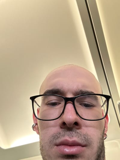 The image presents a close-up, low-angle perspective of a male individual's head and upper face, positioned against a light-colored, uniform background. The individual has a bald or closely-shaved scalp, prominent eyebrows, and visible stubble or short facial hair on the chin and cheeks. He is wearing black-framed eyeglasses. In his left ear, two silver-colored hoop earrings are visible, with a single silver-colored hoop in his right ear. His gaze appears directed slightly downwards or directly at the camera from the low angle. The background is an unadorned, cream-colored surface, likely an interior ceiling or upper wall. A vertical dark line or seam runs along the right edge of the background, potentially a structural element or part of a linear light fixture. No specific geographic location, including the city of Stolbishche, Russia, is discernible from the image content. There are no overt actions or interactions with other subjects.