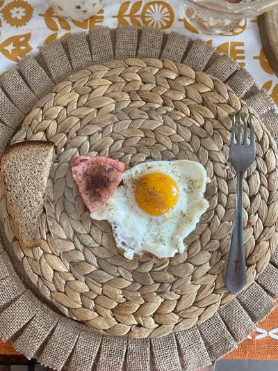 A fried egg with a runny yolk, a slice of dark bread, and a piece of pink ham are arranged on a round, woven placemat with a burlap fringe. A metal fork rests to the right of the plate. The placemat is on a tablecloth with a yellow floral pattern. The scene appears to be a breakfast setting. The location is not specified in the image.