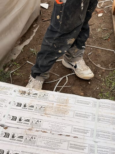 The image shows a close-up view of a person's legs and feet wearing dirty black work pants and white Nike sneakers, standing on muddy ground next to some white plastic sheeting and stacked materials. The stacked materials appear to be boxes or packaging for tiles or similar construction materials, as indicated by printed text and symbols on their surfaces. The ground is composed of dark soil with scattered debris. The context suggests a construction or renovation site. The city of Příbram, Czechia, is mentioned as the location, but no specific landmarks or features in the image confirm this.