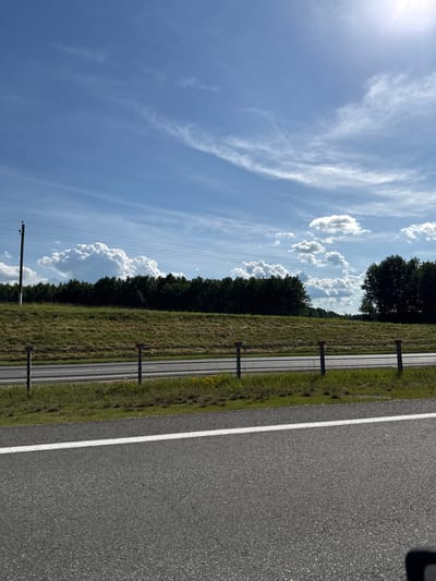This is a landscape photo of a rural road in Belarus. The scene is dominated by a bright blue sky with scattered white clouds, some wispy and others puffy. The sun is visible in the upper left corner, casting a bright glare.
To the right of the sky is a dense line of dark green trees. Separating the trees from the road is a grassy area with some dry, golden-brown patches, suggesting a warm season. Along the edge of the road, there are several dark posts with red tops, which appear to be some form of guardrail or marker.
The road itself is paved and dark gray, with a clear white line marking its edge. The perspective suggests the photo was taken from a slightly elevated position, looking down the road. In the far distance, faint outlines of more trees can be seen. There are also power lines visible in the sky, supported by a utility pole on the left side of the frame.
The overall impression is one of a quiet, sunny day in the countryside. The lighting and the presence of both lush green and dry grass suggest it could be late spring or summer. There is no visible text in the image.