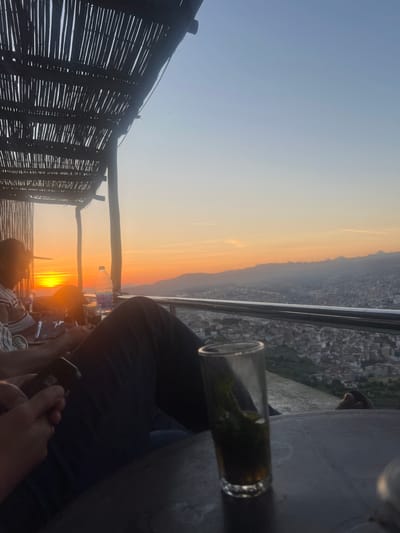 The image depicts an outdoor scene at sunset, captured from a elevated vantage point such as a cafe or terrace overlooking a city. The original image is oriented vertically, but the scene is best understood when rotated 90 degrees counter-clockwise.

In the foreground, a clear glass, likely containing mint tea with visible green leaves, rests on a table to the right. Next to it, a person's legs and a hand holding a smartphone are visible, indicating someone is seated and engaged with their phone. Another person's head and shoulder are partially seen towards the bottom center, facing the sunset. A plastic water bottle also sits on the table. To the left, a dark, rustic woven fence or partition, possibly made of bamboo or reeds, forms a boundary.

The middle ground and background are dominated by a beautiful sunset. The sun is low on the horizon, casting a vibrant orange and yellow light across the sky, which softly transitions to a clear light blue overhead. Silhouetted against the bright sky are distant mountains or hills. Below the horizon, a sprawling city extends across the landscape, with numerous buildings appearing dense and clustered, fading into the atmospheric haze.

The overall scene conveys a sense of relaxation and appreciation for the evening view. The time of day is clearly sunset, with clear weather. Based on the provided context, the location is Tétouan, Morocco, which aligns with the cityscape and the presence of mint tea. No visible text is legible in the image.