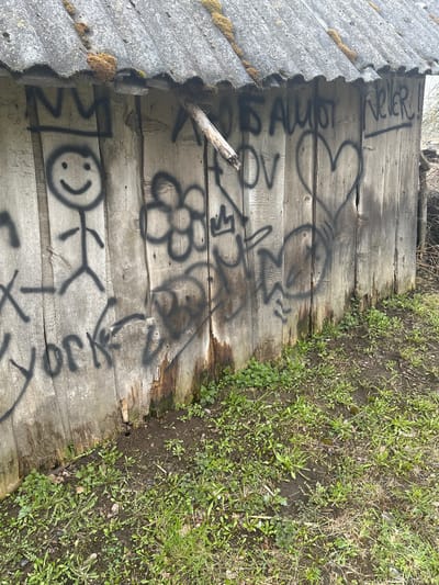 The image depicts a section of an old, weathered wooden fence or wall in Студзёнкаўскі сельскі Савет, Belarus. The wood is grayed and shows signs of age and decay. The surface is covered in black spray-painted graffiti, including hearts, a flower, a stick figure, and words or phrases. Some of the graffiti appears to be faded or incomplete. To the left of the wooden structure is a section of corrugated roofing material with moss growing on it. Adjacent to the right edge of the wooden structure is a patch of grass and various small plants growing in the soil. The overall scene suggests an abandoned or neglected area.