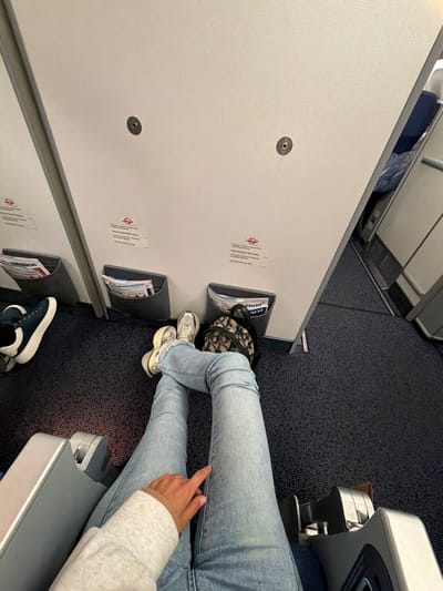 A person seated on an airplane. The person wears jeans and sneakers. A small patterned bag is under the seat in front. Airplane features are visible: seat divider, literature holder, and blue carpet. The image is taken from an overhead perspective, emphasizing the limited legroom.