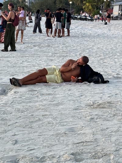The scene is Cenang Beach, Malaysia. A light-skinned adult male, bald with a beard, lies on his left side on light-colored sand. He wears light yellow shorts and dark camouflage-patterned flip-flops. His upper torso is bare. His left hand holds an orange-cased smartphone, with his gaze directed at the device. A black item of clothing or bag serves as a pillow under his head. His right hand rests near his mouth. He is positioned on a light-colored towel. In the mid-ground, several individuals are present: a female with a patterned head covering, wearing a dark top and pants, and a red backpack, stands viewing a mobile device; another female in a floral dress holds a small child dressed in purple. A group of young males stands clustered further back on the sand. The background features resort buildings, palm trees, and multiple parked jet skis. The sand exhibits ripples and footprints.
