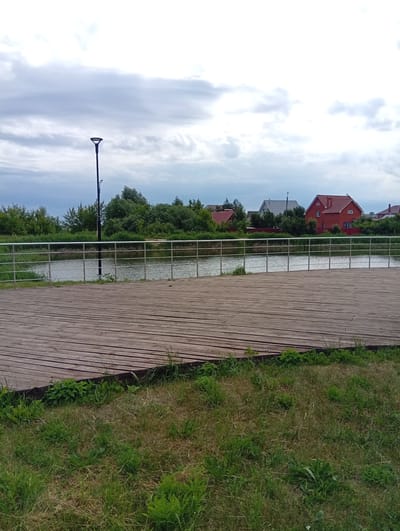 This is a medium shot, eye-level, outdoor, daytime, slightly overcast photo of a wooden boardwalk in Ruzaevka, Russia. The boardwalk, made of light brown wooden planks, takes up the entire lower half of the frame. In the middle ground, a body of water, likely a river or lake, is visible. A metal railing with horizontal bars runs along the edge of the boardwalk, separating it from the water. Beyond the water, a dense line of green trees and bushes forms the background, with a few houses peeking through the foliage. The houses have red tile roofs and some have white or gray walls. A tall, dark lamppost stands on the left side of the frame, near the water's edge. The sky is filled with thick, gray clouds. The overall mood is peaceful and serene.