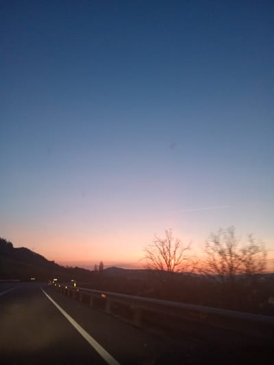 The scene depicts a rural road at either dawn or dusk in the area of Casas del Puerto, Spain. The upper two-thirds of the image show a clear sky transitioning in color from deep blue at the zenith to lighter blue, then purple, and finally orange-pink at the horizon. A faint white contrail is visible horizontally across the lighter blue portion of the sky. The lower third features an asphalt road, viewed from a moving vehicle, with a solid white line marking the visible lane's left edge. To the left, a dark, vegetated hillside with white reflective chevron markers ascends. To the right, a metal guardrail equipped with yellow/orange reflective markers runs parallel to the road, beyond which are blurred silhouettes of bare deciduous trees. No persons or other vehicles are present. The ambient illumination results in the landscape appearing largely in silhouette against the colorful sky.