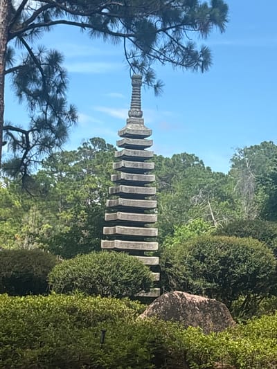 The image captures a stone pagoda in a lush garden setting. The pagoda, constructed from stacked stone layers, is positioned centrally within the frame. It is surrounded by a variety of green bushes and trees, creating a natural and serene atmosphere. A large rock is visible in the foreground, partially obscured by the foliage. The sky above is clear and blue, with some wispy clouds. The scene appears to be an outdoor garden, likely part of a botanical garden or park, given the manicured landscaping and the presence of the pagoda, which suggests a Japanese or East Asian influence. The image was taken in Delray Beach, United States.