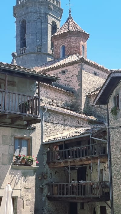 The image shows a view of the town of Rupit, Spain, featuring historic stone buildings and a church bell tower. In the foreground, to the left, is a portion of a stone building with a wooden balcony. The balcony has a railing and a potted plant. Below the balcony is a window with a flower box containing red and pink flowers. To the right of this building are two multi-story wooden balconies with railings, attached to another stone building. These balconies appear weathered and have some potted plants and what look like flags hanging from them. Further in the background, a large stone bell tower with arched openings rises against a clear blue sky. Next to the bell tower is a smaller, octagonal brick structure with a tiled roof, topped with a cross. The overall impression is of an old European town with traditional architecture and a sunny, clear day.