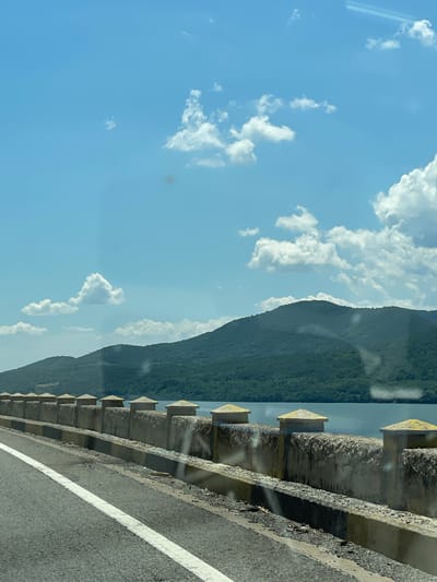 This image presents a daytime scene from a road situated alongside a large body of water, with a backdrop of forested mountains under a clear blue sky. The location is consistent with the Danube River landscape near Drobeta-Turnu Severin, Romania.
In the foreground, a grey asphalt road occupies the lower-left, featuring a worn, solid white line marking the edge of the travel lane. Parallel to the road, a concrete barrier separates it from the water. This barrier consists of a lower curb, a textured concrete wall, and a series of evenly spaced concrete posts topped with light yellow, somewhat conical caps. Debris, including small stones and litter, is visible along the base of the wall. Reflections and smudges, characteristic of a window, are apparent across the entire image, notably in the sky and water areas.
The midground features a calm, blue-grey body of water, presumably the Danube River, extending to the base of a series of lush, green, densely forested mountains.
The background is dominated by the undulating profiles of these mountains, covered in extensive tree canopy. Above the mountains, a bright blue sky contains numerous scattered white cumulus clouds of varying sizes. A small, dark, indistinct object, possibly avian, is visible high in the sky above the mountain range. No identifiable individuals or direct interactions are discernible within the frame.