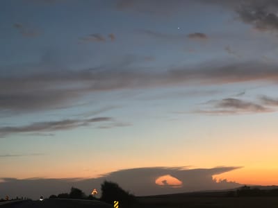 The image captures a twilight scene on a highway leading into Tary, Kazakhstan. The sky is a gradient of deep blue at the top, transitioning to light blue and then a warm orange hue near the horizon, indicating the sun has recently set. Wispy clouds are scattered across the sky, some tinged with pink and orange. A single, bright star is visible in the upper right portion of the sky.

In the foreground, a dark asphalt road curves slightly to the left, suggesting a vehicle's perspective. The road is bordered by dark, silhouetted trees and vegetation. Two road signs are visible: one yellow triangular sign with a black symbol of three vertical lines stacked on a horizontal line, indicating a hazard or uneven road surface, is positioned on the right side of the road. Another sign, partially visible on the left, is also yellow and triangular with a black symbol. The headlights of at least two vehicles are faintly visible on the road ahead.

The landscape in the distance is also silhouetted against the brightly lit sky, with the faint outline of hills or mountains visible on the horizon. A peculiar cloud formation with a distinct hole is visible in the middle distance, casting a shadow on the landscape below. The overall atmosphere is serene and captures the transition from day to night in the rural outskirts of Tary.