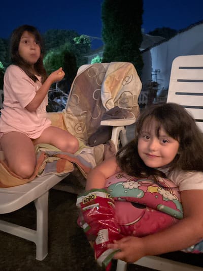 Two young girls are seated on plastic chairs outdoors at dusk. The girl in the foreground, on the left, is wearing a pink t-shirt and looking towards the camera with a curious expression. She is lying on her back. The girl in the background, on the right, is leaning back on her chair and holding a pillow with a cartoon unicorn design and a bag of snacks. She is looking at the camera with a slight smile. The background shows dark foliage and a building with exterior lights, suggesting a residential setting. The overall mood appears relaxed and familial. The time of day is likely evening, as indicated by the dark blue sky. Visible text on the snack bag reads "LOTTO".