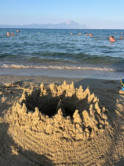 The image depicts a beach scene in Toroni Municipal Unit, Greece. The foreground is dominated by a close-up view of a hole dug in the sand, with sunlight casting shadows that highlight the granular texture of the sand and the irregular edges of the hole. In the background, the calm, blue water of the sea meets the shore with gentle waves. Several people are visible in the water, swimming and enjoying the sea. The water is clear, allowing glimpses of the sandy seabed. Beyond the water, a hazy mountain range can be seen under a clear, light blue sky. The presence of buoys in the water suggests a designated swimming area. The lighting indicates it is likely late afternoon or early evening, with the sun casting long shadows. The overall atmosphere is serene and recreational. No visible text is present in the image.