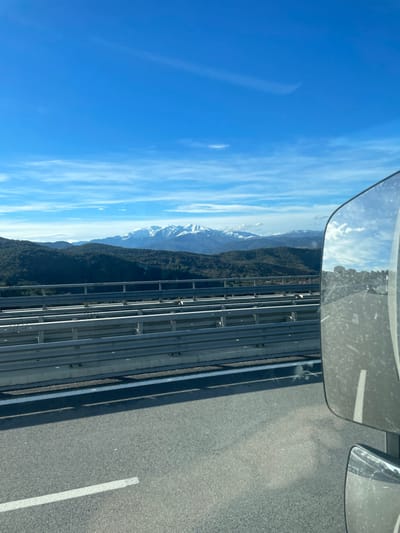A view from a high bridge shows a road with a guardrail and a distant mountain range. The peaks are dusted with snow against a blue sky. A rearview mirror is visible in the upper left corner, reflecting the sky. The road surface is gray, marked with white lines, suggesting a highway. The scene implies travel, with nature's grandeur as a backdrop.