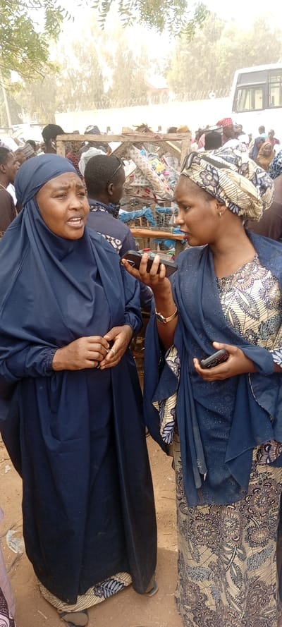 An outdoor scene in Sani Mai Nagge, Nigeria, depicts two adult females in the foreground, engaged in an interaction amidst a crowded environment. The woman on the left, facing slightly right, wears a dark blue abaya with a matching head covering, revealing her face. Her mouth is open, suggesting speech. Her hands are clasped at waist level, with a silver-colored ring visible on her right ring finger. The woman on the right, facing slightly left towards the first woman, wears a patterned dress in beige and blue tones, a matching patterned headwrap, and a translucent dark blue overlay garment. She holds a black smartphone horizontally in her right hand, positioned near the first woman's face, with the screen oriented towards her. A gold-colored wristwatch is on her left wrist.
In the midground, a dense crowd of people is visible. Behind them, several rudimentary market stalls or structures made of wooden frames and wire mesh hold assorted goods, including textiles and plastic items. The ground is unpaved, appearing as dirt or sand. In the background, green foliage from trees is visible on the left, and a partial view of a white bus or large van is seen on the right. A fence, possibly topped with barbed or razor wire, separates the immediate area from a distant building. The overall atmosphere is bright and appears dusty or hazy.
