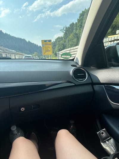 The image shows the interior of a car, looking out through the windshield. The perspective is from the passenger seat, with a person's legs visible in the lower right portion of the frame. Outside the car, a bright, sunny day is evident with a blue sky and some clouds. Green trees and hills form the backdrop. A yellow road sign with blue text and arrows is visible through the windshield, indicating directions.
Inside the car, the dashboard is visible, with an air vent and the word "AIRBAG" printed on it. A green sticker with white text is affixed to the windshield. To the right, a black handbag with a zipper is partially visible. A bottle of Volvic Vitamin Water is on the floor in the passenger footwell. The overall impression is of a road trip or travel on a pleasant day in Gernsbach, Germany.