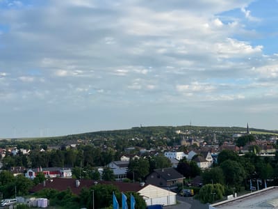 The image shows a panoramic view of Alzey, Germany, under a partially cloudy sky. In the foreground, there are rooftops of houses and commercial buildings, with trees interspersed throughout the scene. To the left, several white wind turbines are visible on a distant green hill. The middle ground is filled with a dense cluster of buildings, including residential houses and larger structures that appear to be commercial or municipal. A church steeple is prominent among the buildings. The background features rolling green hills, also dotted with wind turbines and some smaller structures. The lighting suggests it is daytime, possibly late afternoon or early morning, with soft shadows. There are no visible people or animals in the image.