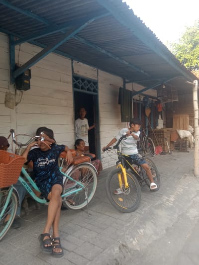 The image depicts an outdoor scene in what appears to be a village setting in Jumput, Indonesia. The foreground features two children with bicycles. A young girl, wearing a black t-shirt with a blue pattern and black shorts, is seated on a light blue bicycle. She is wearing dark sandals. Her legs are crossed, and her left hand rests on the handlebars. To her right, a younger boy in a striped t-shirt and patterned shorts is positioned on a yellow and black mountain bike, looking to his right. He is wearing white slides.
Behind the children, a light-colored wooden house with a corrugated metal roof supported by blue beams is visible. An open doorway reveals a man in a white t-shirt standing inside. To the right of the doorway, an older woman is seated and smiling, wearing a patterned skirt and shirt. Further back, there are more bicycles, birdcages hanging from the roof, and stacks of what appear to be cardboard or wood. The ground is paved with interlocking gray bricks. The overall impression is of a casual, everyday scene in a residential area.