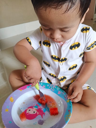In Cibungbulang, Indonesia, a young child wearing a white t-shirt with a Batman logo pattern is sitting on a tiled floor. The child is looking down at a colorful plate with a mermaid design, attempting to eat watermelon with a fork. The plate contains several pieces of watermelon and some juice. The scene appears to be an indoor setting, possibly a home.