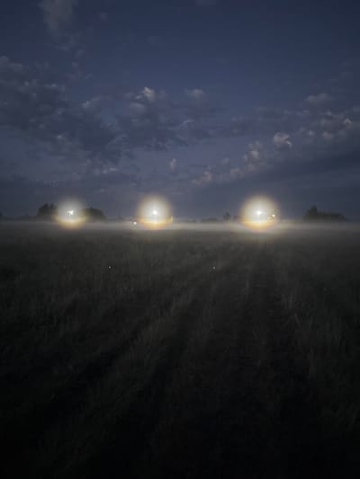 The image depicts a dark, foggy field at night. Three bright, glowing orbs of light are visible in the middle ground, appearing to be from vehicle headlights or stationary lights. These lights are surrounded by a dense mist, obscuring the details of the landscape and any potential subjects. The foreground consists of dark grass. The sky above is a deep blue-grey, with scattered clouds. There is no discernible indication of the city of Ворошиловка, Kazakhstan.