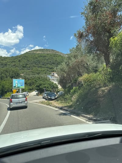 The image captures a sunny day on a road in Vrijesno, Montenegro, with a lush, green mountain backdrop. In the foreground, a white car's dashboard and windshield are visible, implying the photo was taken from inside a vehicle. A silver car is driving ahead on the asphalt road, which has white lane markings. To the right of the road, a dense green hillside is covered in trees and shrubs, with a few buildings, including a white one with balconies, nestled among them. A billboard advertising "KEFIR" is positioned on the left side of the road. Several other cars are parked or driving in the vicinity. A person in a black top and shorts is standing near the parked cars on the right side of the road. The sky is blue with scattered white clouds.