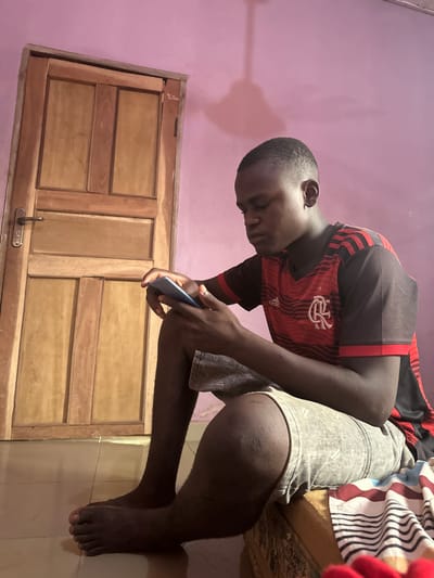 An image captured in Ozalla, Nigeria, depicts a male, approximately 18-25 years old, seated indoors. He has dark skin and a short, faded haircut. The individual is focused on a light-colored smartphone held horizontally in both hands. He wears a red and black horizontally striped short-sleeved t-shirt with an Adidas logo and a CR Flamengo club crest on the chest, along with light grey denim shorts. He is barefoot.
The subject is positioned with his left foot flat on a light-colored tiled floor and his right thigh resting on a low, firm surface, partially covered by a multi-striped fabric (red, white, blue, black). In the background, a light-brown wooden door with a metallic handle is visible to the left. The wall behind the subject and the door is painted a solid pale pink or light purple. A distinct shadow, consistent with a ceiling fan or light fixture, is cast on the upper section of the pink wall.