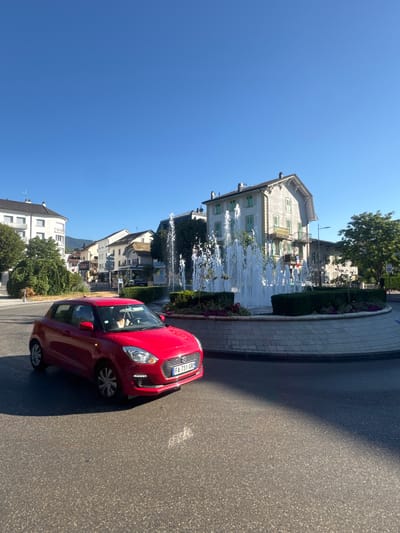 A red Suzuki Swift is turning through a roundabout in Divonne-les-Bains, France. A woman is visible behind the steering wheel. The roundabout features a large, multi-tiered fountain with water spraying upwards, surrounded by manicured green hedges and flowering plants. In the background, buildings with light-colored facades and green shutters line a street. Trees with lush green foliage are present, and the sky is clear and blue. The image is captured on a sunny day, indicated by the strong shadows cast on the road. The license plate of the car is visible and reads "FA 731 GR".