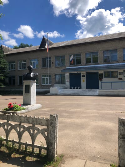 A bust of a person stands on a pedestal in front of a brick school building with a corrugated roof in Izdeshkovo, Russia. A Russian flag flies from a pole on the roof. The building has several windows and two blue doors. The area in front of the building is paved, and a decorative concrete fence runs across the foreground. Red flowers are planted around the base of the bust. The sky is blue with white clouds.