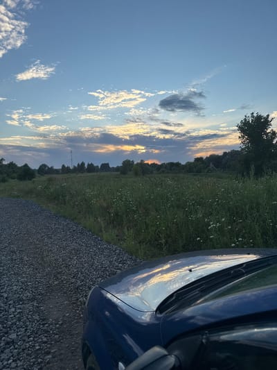 An outdoor, slightly elevated shot captures a twilight scene in the rural outskirts of Kovrigino, Russia. The foreground is dominated by the hood and windshield of a dark blue car, reflecting the sky. To the left of the car is a gravel road, with a grassy field of wildflowers stretching into the distance on the right. The field is bordered by a line of trees and brush. A distant cell tower is visible in the center background. The sky is a mix of blue and wispy white and grey clouds, with the setting sun casting a warm, golden light.