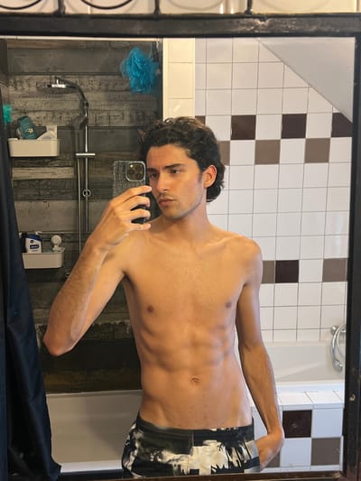 A young man with dark, curly hair is taking a mirror selfie in a bathroom. He is shirtless and wearing patterned shorts. In the background, a showerhead and toiletries are visible on a tiled wall. The bathroom also features a white bathtub and checkered tiles on another wall. The man's physique is lean with visible abdominal muscles. The scene suggests a private, personal moment.
