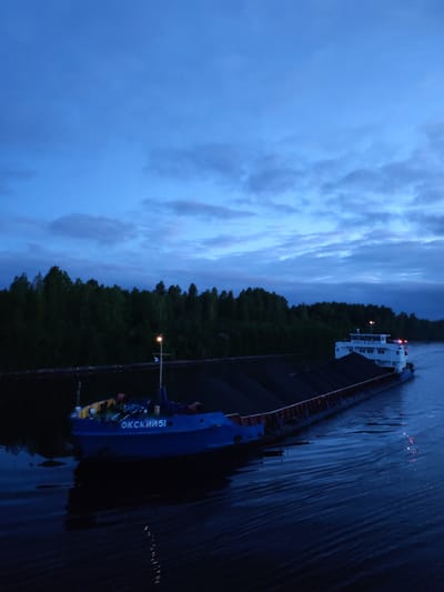 The image shows a barge, likely in the city of Annensky Most, Russia, on a body of water at dusk or twilight. The sky is a mix of blue and gray clouds. The barge is loaded with what appears to be a dark cargo, possibly coal. The barge is composed of two sections, with a blue tugboat at the front, and a larger freighter section at the rear. The tugboat has "OKSKII 51" written on the side in Cyrillic script. The freighter section is white on the top, with Cyrillic script along the top. The water is dark and rippled, reflecting the sky. The background consists of a dense forest along the shore.