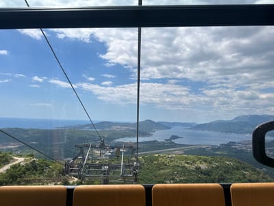 The image is taken from inside a cable car, looking out over a landscape in Mirac, Montenegro. The cable car's frame and seats are visible in the foreground. Through the glass, there is a view of a coastal region with water, mountains, and green vegetation. Visible are infrastructure and a road along with the surrounding landscape. The sky is blue with scattered clouds.