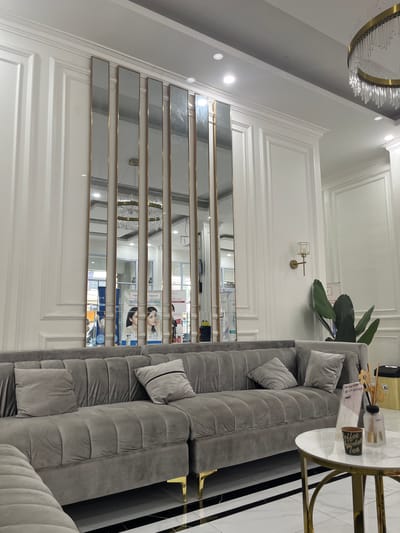 This image captures an interior scene, likely a waiting area or reception space, with a prominent grey, channel-tufted sectional sofa dominating the foreground. Two throw pillows, one plain grey and another grey with a black stripe, rest on the sofa. A round, white marble-topped coffee table with a gold-colored base sits to the right of the sofa, holding a small potted plant and a glass diffuser with wooden reeds.
The background features a wall adorned with elaborate white molding and several tall, vertical mirrors with gold frames, which reflect a chandelier and some posters or advertisements. To the right of the mirrors, a wall sconce with a crystal shade is mounted. A large-leafed plant is positioned in the corner between the sofa and the wall. The floor is tiled in a black and white pattern. The overall aesthetic suggests a modern, clean, and possibly upscale environment.
The location of this scene is a clinic or spa in Dumai City, Indonesia. The posters reflected in the mirrors appear to advertise cosmetic or dermatological services, featuring images of women and text in what seems to be Bahasa Indonesia. The ambiance is calm and inviting.