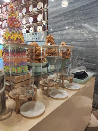 The image shows a dessert buffet, likely in Ürümqi, China, given the context of the prompt. In the foreground, there are several clear glass dispensers with wooden lids, standing on wooden stands. The dispensers are empty, except for a small amount of liquid at the bottom of one. White plates are arranged around the dispensers. To the left, a tall, cone-shaped decoration resembling a Christmas tree is covered in colorful, candy-like elements. Behind the dispensers, a large bowl of golden-brown, spiral-shaped fried pastries is visible, with more pastries stacked in clear cylindrical containers. To the right of the pastries, a light blue tray holds a stack of black disposable masks. The background features a wall with a marble-like pattern and shelves displaying circular objects. A commercial disinfection cabinet is also visible in the background. The lighting suggests an indoor setting, possibly a hotel or restaurant. There are no people visible in the image.