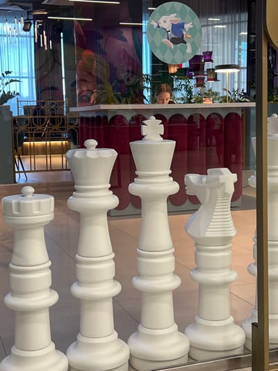This media file captures a display of oversized white chess pieces, possibly a decorative installation. The pieces are arranged vertically, suggesting a layered presentation. The closest chess piece appears to be a queen or king, characterized by its elaborate crown-like top. Below it, another piece, possibly a queen, is visible, followed by a rook. The furthest visible piece is a knight, distinguished by its horse-head design.

The background is complex and reflective, showing parts of an interior environment. There are glimpses of furniture, lighting fixtures, and plant life, indicating an indoor setting such as a shop, gallery, or public space. A circular decal featuring a cartoon rabbit in a blue jacket is visible on the left, adding a playful or whimsical element to the scene. Reflections in the glass casing reveal a person's face and upper body, suggesting the viewer is observing the chess pieces through a glass barrier. The lighting appears to be artificial, and there are no indicators of the time of day or weather conditions. The overall impression is one of a visually striking and artistic arrangement of chess pieces within a contemporary interior. The location context of Borisova Griva, Russia, is provided but not visually discernible from the image itself.