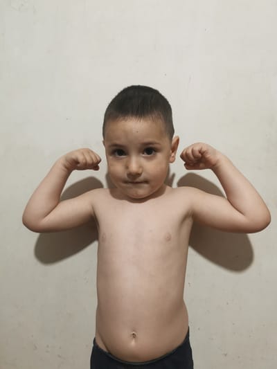 A young boy is posing in front of a textured, light-colored wall, likely indoors due to the lighting and plain background. He is shirtless and wearing dark-colored shorts. The boy has short, dark hair and is looking directly at the camera with a neutral expression. His arms are bent at the elbows, with his fists clenched and held up, as if he is flexing his muscles. Shadows cast by his arms are visible on the wall behind him. The lighting appears to be artificial, suggesting the photo was taken inside. There is no visible text in the image. The setting is identified as Carbuna, Moldova.