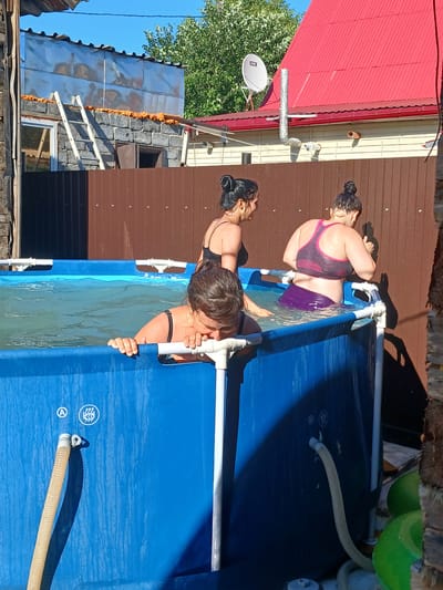 In an outdoor setting, two women are enjoying a swim in a blue, rectangular above-ground pool. The water is clear and inviting, reflecting the bright sunlight. One woman, in a purple bikini, is partially submerged, with her head tilted back and eyes closed. The other woman, wearing a black swimsuit, is leaning over the edge of the pool, appearing to reach for something in the water. Both women have their hair pulled back in buns.
The scene appears to be a backyard in Chik, Russia, with hints of residential structures in the background. A red-tiled roof and a brown wooden fence are visible, along with some greenery from trees. The time of day is clearly daytime, with strong sunlight casting shadows. The weather seems warm and pleasant, suitable for swimming.
Notable details include the inflatable nature of the pool, indicated by its white frame, and a flexible hose connected to a filter system at the bottom of the pool. There are also some circular symbols with wave-like patterns and a letter 'A' printed on the pool liner. The overall atmosphere suggests a relaxed and enjoyable summer activity.