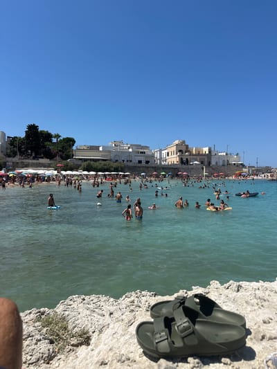 The image captures a crowded beach scene on a clear, sunny day at Santa Maria al Bagno, a coastal frazione of Nardò, Italy. In the immediate foreground, a pair of dark green, two-strap slide sandals rests on light-colored, porous rock formations, with a bare human arm and leg partially visible to the left. The midground is dominated by a clear, turquoise-blue body of water teeming with numerous individuals. These people are engaged in various actions including swimming, standing in the water, and floating on inflatable devices, specifically a blue mat and a yellow ring. The water extends to a sandy beach in the background, densely populated with sunbathers. A large quantity of white and some red beach umbrellas are arranged in rows across the sand. Beyond the beach, a retaining wall supports a landscape featuring multiple light-colored, multi-story buildings typical of Mediterranean architecture, set against a backdrop of green trees. Prominent structures include a large white building with an outdoor seating area and, to its right, a distinct tan building with arched windows. On the far right, a pier or jetty extends into the water. The sky is a uniform, cloudless bright blue.