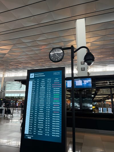 An indoor shot captures a digital flight information board at an airport. The board, a large rectangular screen, displays a list of flights with columns for airline, flight number, destination, check-in counter, and status. The "General Check-in Terminal 3 Domestic" title is visible at the top of the board. A clock with black hands on a white face is mounted above the information board. A security camera is attached to a black pole extending upwards from behind the information board. The airport environment features a modern ceiling with geometric patterns and tiled flooring. Several blurred figures of people can be seen in the background. The scene is lit by overhead artificial lighting.