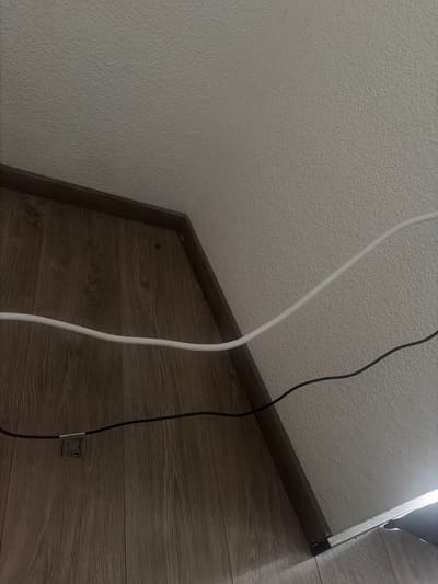 The image depicts a section of a room, showing a wall meeting a corner where it transitions into a structure with a wooden-patterned surface. Two electrical cords, one white and one black, run along the wall and the wooden surface. The wall has a textured surface, possibly stucco or a similar finish. The wooden surface appears to be laminate or veneer with a wood grain pattern. A small tag is affixed to the black cord. The location is unknown, and there's no information to suggest it's in Germersheim, Germany.