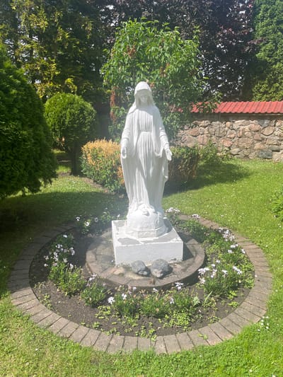 The image shows a white statue of Mary in a garden. The statue is standing on a pedestal surrounded by a circular flower bed with small white and purple flowers and two small hedgehog figures. The flower bed is bordered by a brick pathway, and behind it is a grassy area. In the background, there are several evergreen trees, a stone wall with a red tiled roof, and more trees. The sun is shining, casting shadows on the grass. The scene appears to be outdoors in a garden or a park. It is not possible to determine the exact location within Гервяцкі сельскі Савет, Belarus from the image alone.