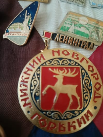 The image displays a collection of Soviet-era commemorative badges. In the foreground, a large, circular gold-colored medal with red and blue enamel is prominently featured. The medal has a depiction of a red shield with a golden stag in the center. Inscriptions in red Cyrillic letters encircle the stag. To the left of the medal, a smaller, triangular badge with an image of the Statue of Liberty and text in red Cyrillic reads "АРХАНГЕЛЬСК" (Arkhangelsk). Above and to the right of the medal, a rectangular badge with a depiction of a building and text in green Cyrillic reads "КАЗАНИ" (Kazan). Below this, another rectangular badge with text in black and gold Cyrillic reads "ЛЕНИНГРАД" (Leningrad). The badges are arranged on a surface that appears to be fabric, with sections of brown and white fabric visible. The lighting suggests an indoor environment. There are no people or actions depicted in the image. The location is not specified as Verkhnyaya Storona, Russia.