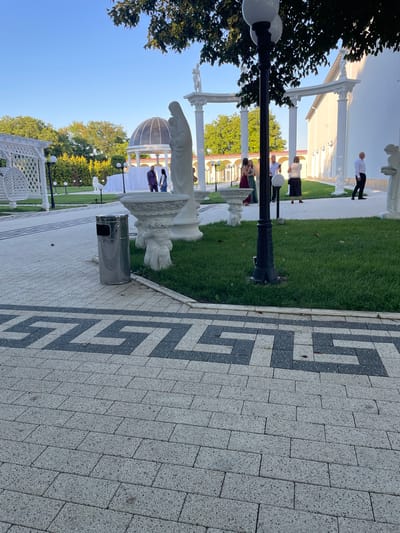 In Ratuș, Moldova, a wedding venue or similar event space is depicted. The foreground is dominated by a paved walkway with a Greek key pattern in dark gray and beige. To the left, a tall, stainless steel trash receptacle stands on the walkway. Beyond the walkway is a meticulously maintained lawn with several white marble statues. One statue, of a woman holding a child, is prominent in the mid-ground. In the background, a white building with classical architectural elements, including columns and arches, is visible. Several people are scattered across the lawn and near the building. A group of three women in formal dresses and two men in suits are gathered near a draped table and a domed structure. Another man in a white shirt and dark pants walks away from the camera on the right side of the image. The sky is clear and blue, suggesting it is daytime. The overall scene conveys an atmosphere of elegance and celebration.