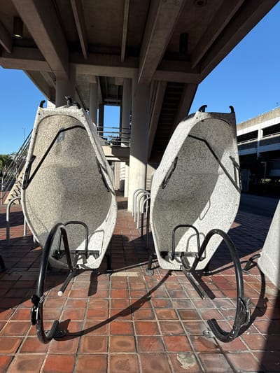 Two futuristic, white, speckled bike racks sit on a red brick walkway. Their black metal frames cast long shadows. Above, concrete architecture with visible stairs and pipes creates a layered scene, punctuated by a patch of bright blue sky. A traditional metal bike rack stands behind the unique ones.