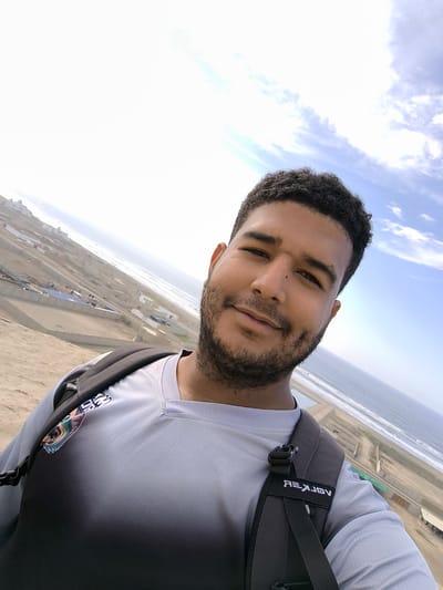 A man wearing a gray and black shirt with a black backpack is taking a selfie in Huanchaco, Peru. In the background are sand dunes, the ocean, and buildings. The sky is partly cloudy.