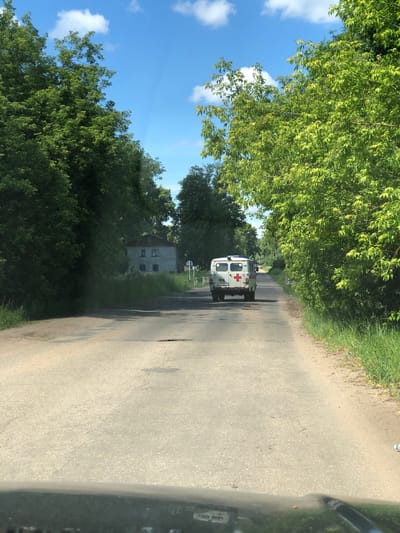 An aerial view from a vehicle's perspective shows an asphalt road in Izdeshkovo, Russia, on a clear, sunny day with scattered clouds. Dense green trees line both sides of the road, creating a canopy effect. In the distance, an ambulance with a red cross emblem is visible, traveling away from the camera. A white sign with a red circle and a diagonal line is posted on the right side of the road. On the left side of the road, a light-colored, two-story building with a dark roof and several windows is partially obscured by trees. The road surface exhibits signs of wear, with visible patches and a pothole. The foreground shows the edge of the vehicle's hood, suggesting the image was taken from the driver's or front passenger's seat.