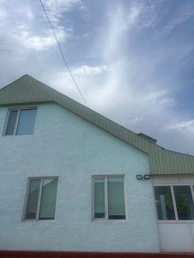 The image shows the exterior of a light blue, two-story building with a green corrugated metal roof. The building has several windows; a tall, narrow window on the upper left, and two rectangular windows on the lower level. To the right of the lower windows is a white-framed glass enclosed entryway or sunroom. Two black outdoor lights are mounted on the blue wall above the lower windows. The building appears to be in a residential or commercial area, with a hint of greenery visible through the glass entryway. The sky above is cloudy with some patches of blue. A thin black wire extends diagonally across the upper portion of the image, from the upper left towards the right. The building is situated in the city of Üshara, Kazakhstan, as indicated by the context.