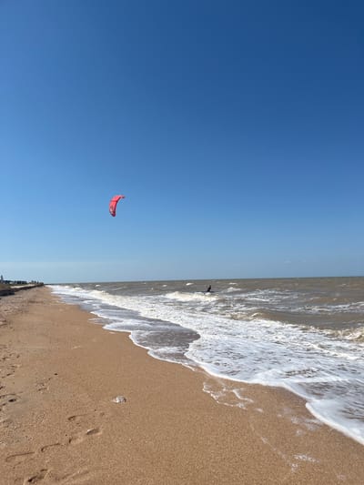 A bright, sunny day at the beach in Dolzhanskaya, Russia. The scene is dominated by a clear blue sky and a wide expanse of tan sand leading to the choppy, brown water. In the distance, a person is kitesurfing, their red kite clearly visible against the sky. Waves are breaking on the shore, leaving white foam on the sand. Footprints are visible in the sand, suggesting recent activity. A few cars are parked along the edge of the beach, near a slightly eroded embankment. The overall impression is one of an active, outdoor recreation area on a beautiful day.