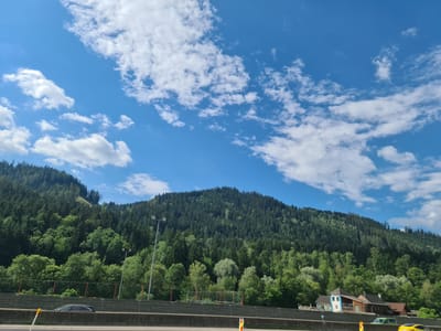 The image shows a scenic view of a forested mountain slope under a blue sky dotted with white clouds. In the foreground, a highway barrier separates the road from the mountain. Several vehicles are visible on the highway, including a dark-colored car and a yellow car. A few buildings, appearing to be houses, are nestled at the base of the mountain on the right side of the image. The overall scene suggests a natural landscape near a populated area.